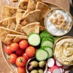 A tan colored round platter filled with pita chips, cherry tomatoes, olives, cucumber, tzatziki and garlic dip.