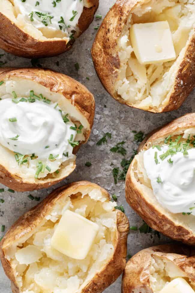 Six baked potatoes topped with sour cream and chives on a baking sheet.