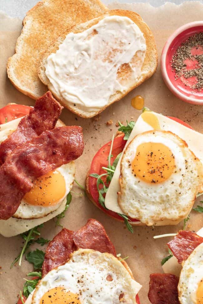 Four fried egg sandwiches with eggs, turkey bacon, cheese, arugula and tomato sit on a piece of parchment paper.
