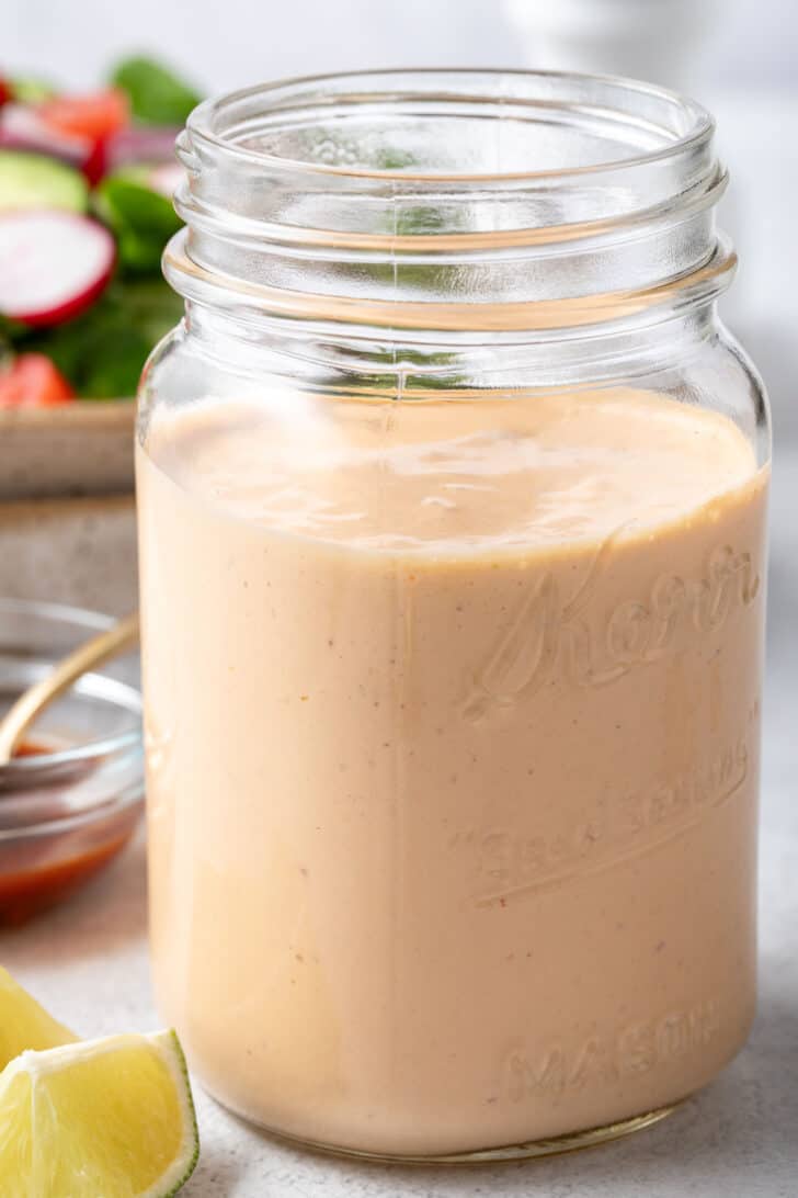 A clear mason jar filled with Chipotle Ranch Dressing. A green salad in a bowl sits behind the jar.