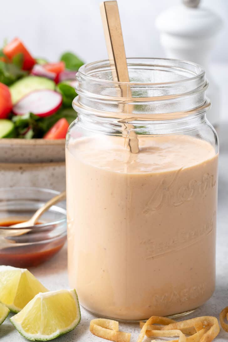 A clear glass mason jar filled with Chipotle Ranch Salad Dressing. A bowl filled with salad sits next to the jar.
