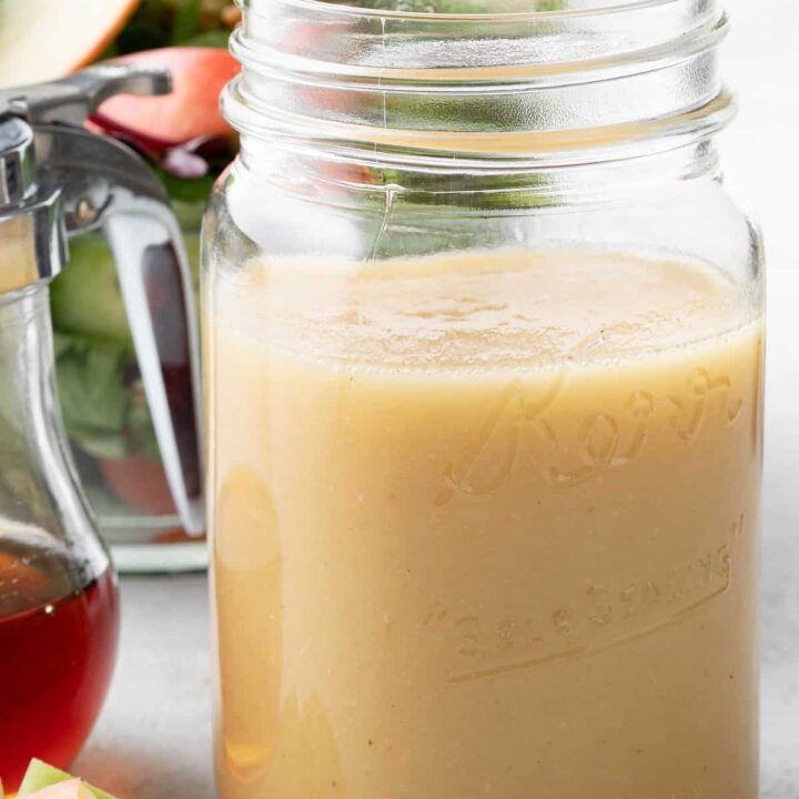 A clear mason jar filled with Maple Vinaigrette. A salad in a clear bowl sits next to the jar.