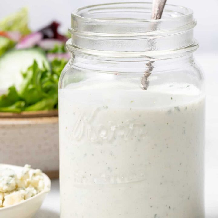 A clear glass mason jar filled with Blue Cheese Ranch Salad Dressing. A tan bowl filled with salad sits behind the jar.