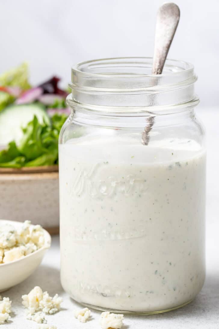 A clear glass mason jar filled with Blue Cheese Ranch Dressing. A tan colored bowl filled with salad sits next to the jar.