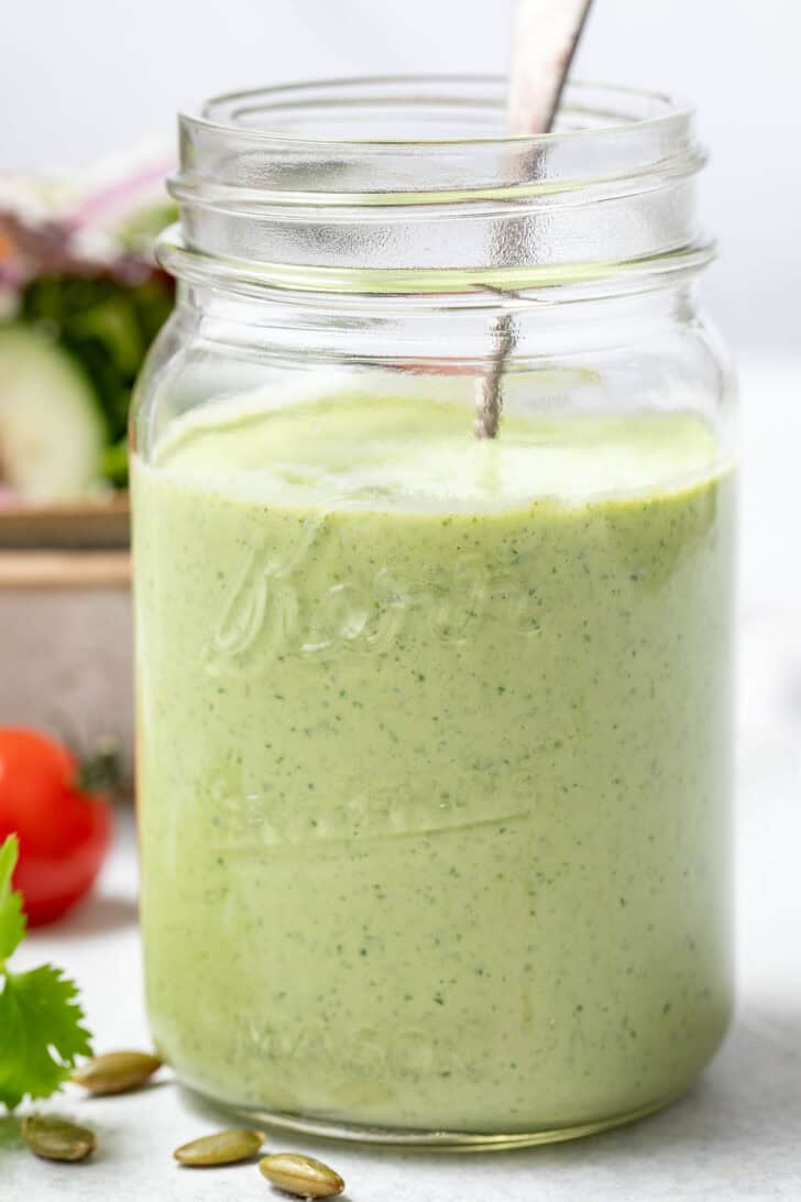 A clear mason jar filled with Creamy Cilantro Salad Dressing. A tan bowl filled with salad sits behind the jar.
