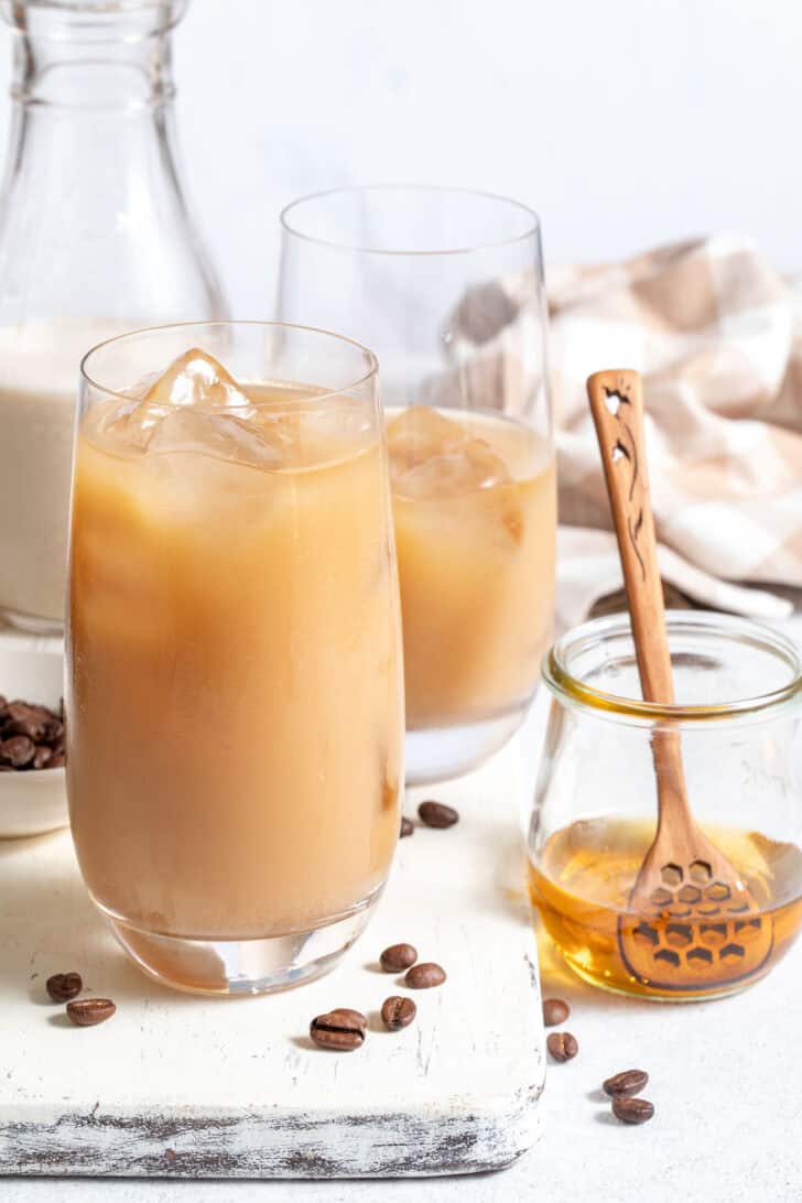 Two clear glasses filled with a chilled drink. A bottle of oat milk and a small jar of honey sit next to the glasses.