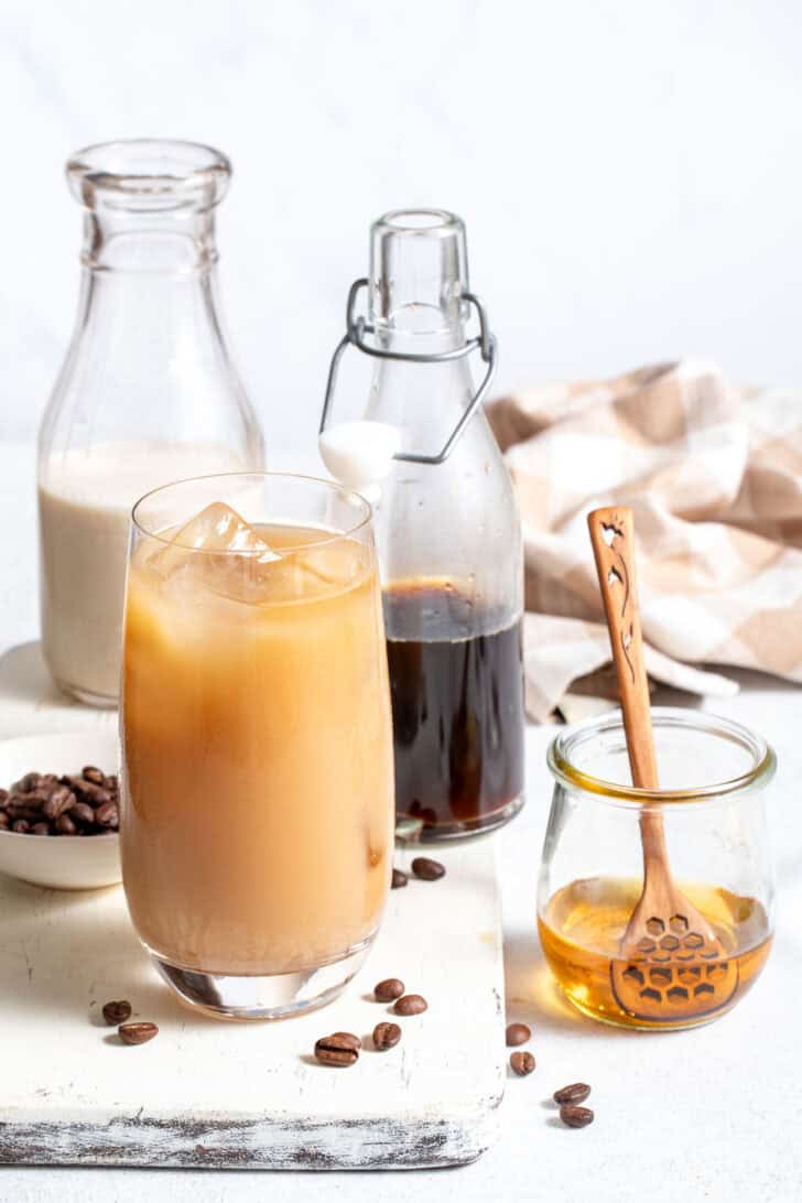 A clear glass filled with Iced Coffee Flavor Water. A bottle of coffee, a bottle of oat milk and a small jar of honey sit next to the glass.