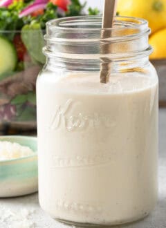 A clear glass mason jar filled with Creamy Lemon Salad Dressing. A clear glass bowl filled with mixed greens sits behind the jar.