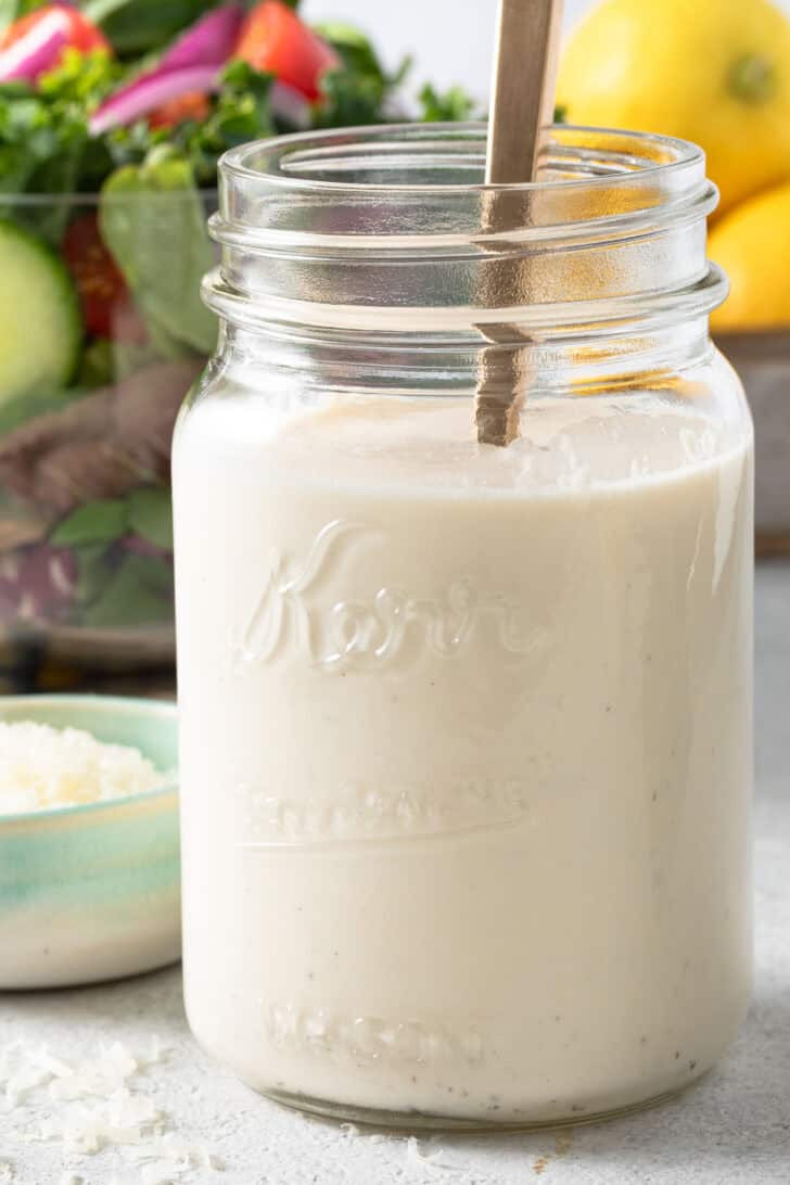 A clear mason jar filled with Creamy Lemon Salad Dressing. A clear bowl filled with mixed green sits behind the jar.