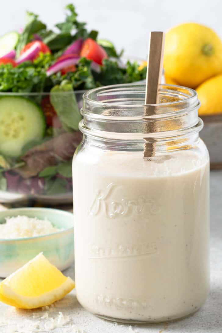 A clear mason jar filled with creamy lemon dressing. A glass bowl filled with mixed greens sits behind the jar.
