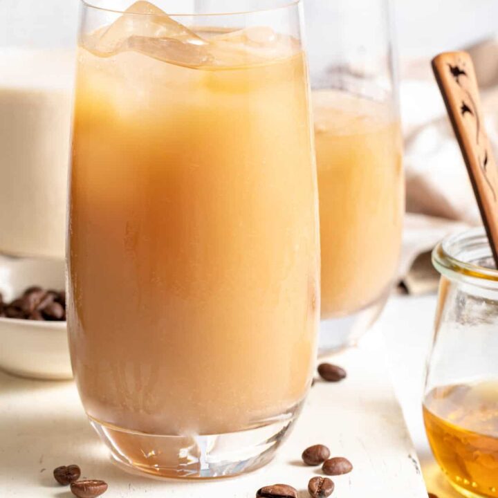 Two clear glasses filled with Iced Coffee Flavored Water. A bottle of oat milk and small jar of honey sit next to the glasses.