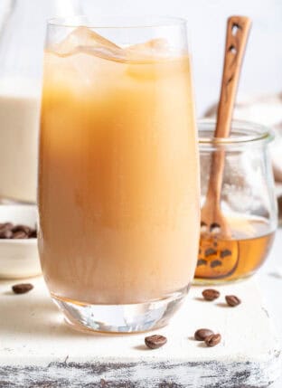 A clear glass filled with Iced Coffee Flavored Water. A bottle of oat milk and small jar of honey sit next to the glass.
