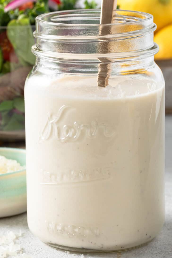 A clear mason jar filled with creamy vinaigrette. A clear glass bowl filled with mixed greens sits behind the jar.