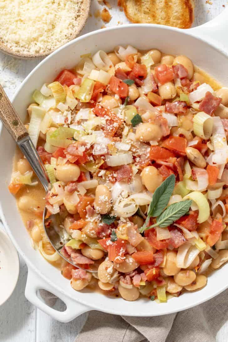 A white skillet filled with canned butter beans, turkey bacon, leeks, garlic and tomatoes for butter beans recipe.