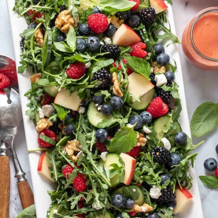 A white platter filled with Arugula Berry Salad. A pair of salad servers and salad plates sit next to the platter.