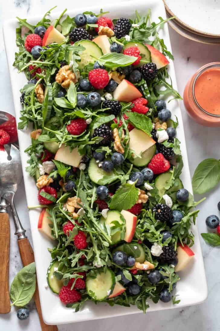 A white platter filled with Arugula Berry Salad. A pair of salad servers and salad plates sit next to the platter.