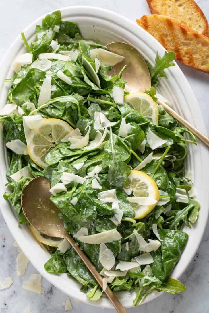 A white oval platter filled with mixed greens. Golden colored spoons rest in the platter.