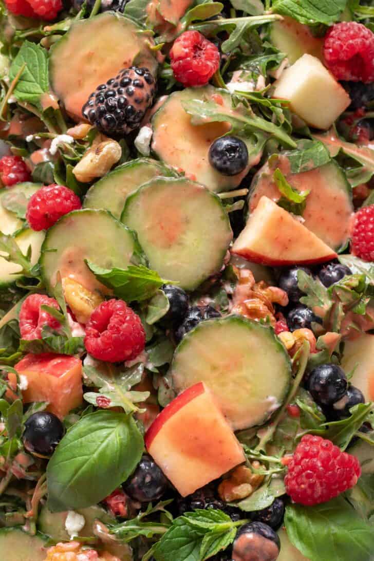 A close up of Arugula Berry Salad - a mix of arugula, cucumber slices, chopped apple, raspberries, blueberries, blackberries, walnuts and fresh basil and mint.