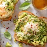 Three Pesto Eggs Avocado Toast on a white countertop. A small glass bowl with honey sits next to the toast.