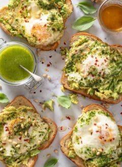 Four pieces of Avocado Toast with Pesto Eggs on a marble board. A glass cup filled with honey sits next to the toas