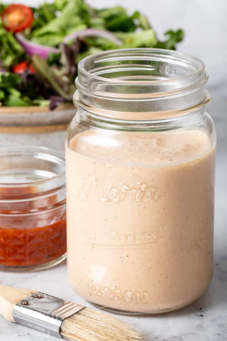 A clear glass mason jar filled with BB! ranch dressing. A tan bowl filled with salad sits next to the jar.