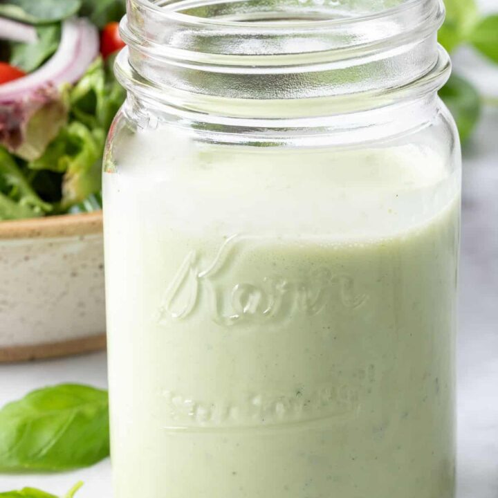 A clear glass mason jar filled with Basil Buttermilk Dressing. A tan bowl filled with salad sits next to the jar.