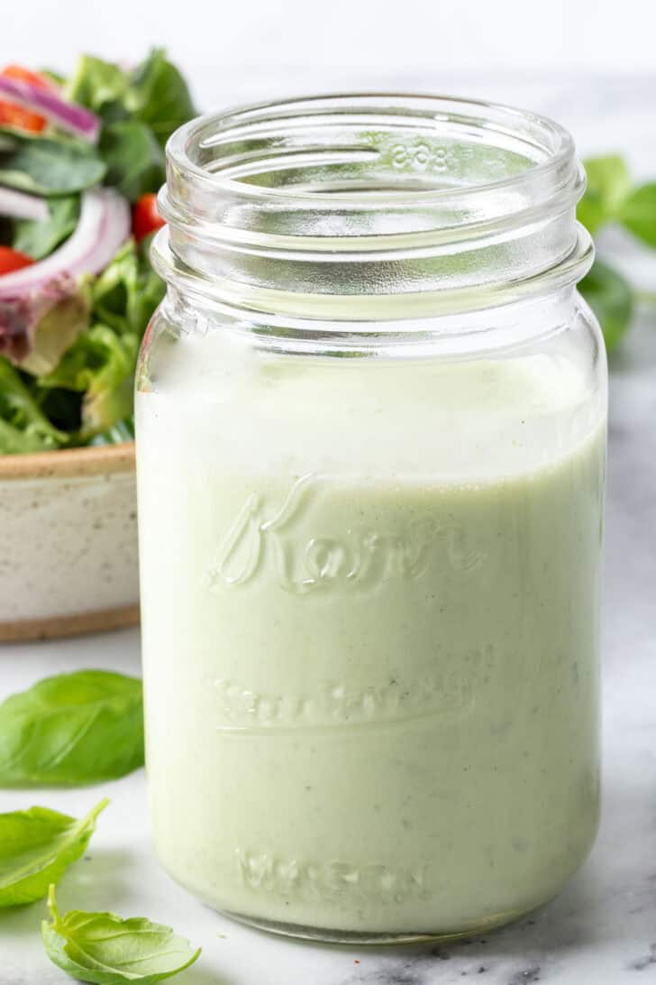 A clear glass mason jar filled with Creamy Basil Salad Dressing. A tan colored bowl filled with salad sits next to the jar.