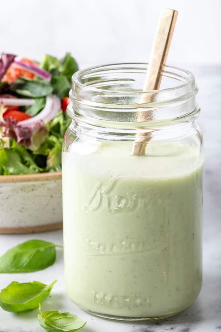 A clear glass mason jar filled with green sauce. A tan colored bowl filled with salad sits next to the jar.