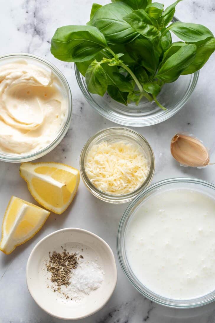 Small clear glass mixing bowls filled with cheese, mayonnaise and fresh herbs sit on a marble cutting board.