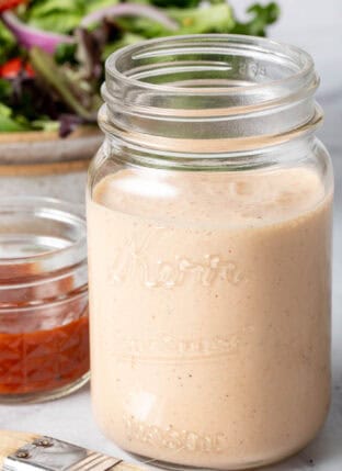 A clear mason jar filled with BBQ Ranch Dressing. A tan bowl filled with salad sits next to the jar.