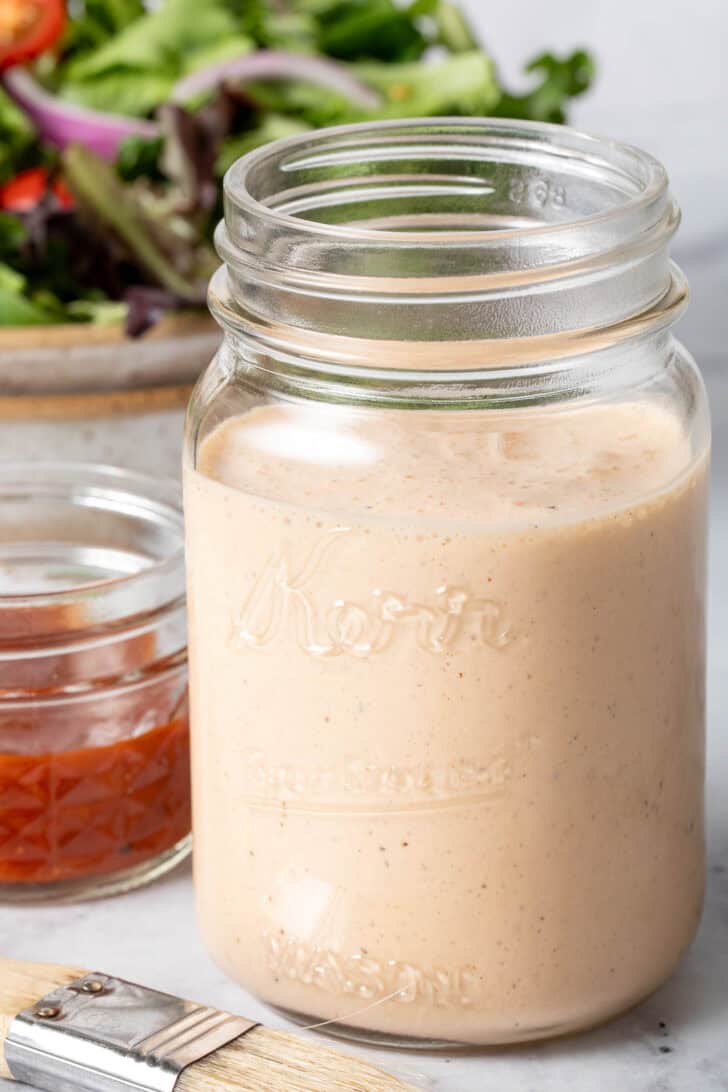 A clear mason jar filled with BBQ Ranch Dressing. A tan colored bowl filled with salad sits next to the jar.