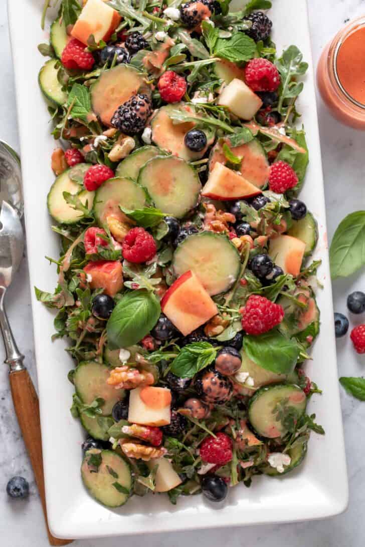 A white platter filled with greens and fruit. A pair of servers sits next to the platter.