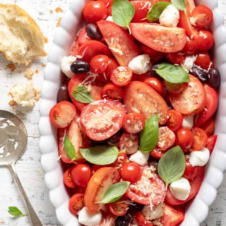 A white oval platter filled with sliced Roma and cherry tomatoes, mozzarella balls, kalamata olives, and basil leaves. A tomato server rests next to the platter.