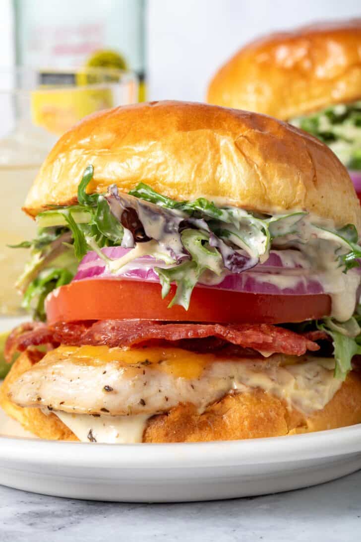 A grilled chicken sandwich with tomato, lettuce, red onion and bacon on a white plate.