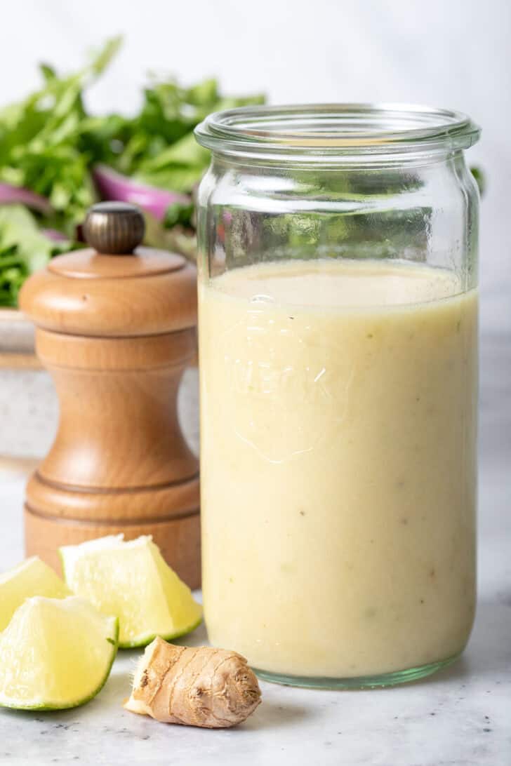 A clear glass mason jar filled with Lime Ginger Vinaigrette. A pepper mill and a bowl of greens sits next to the jar.