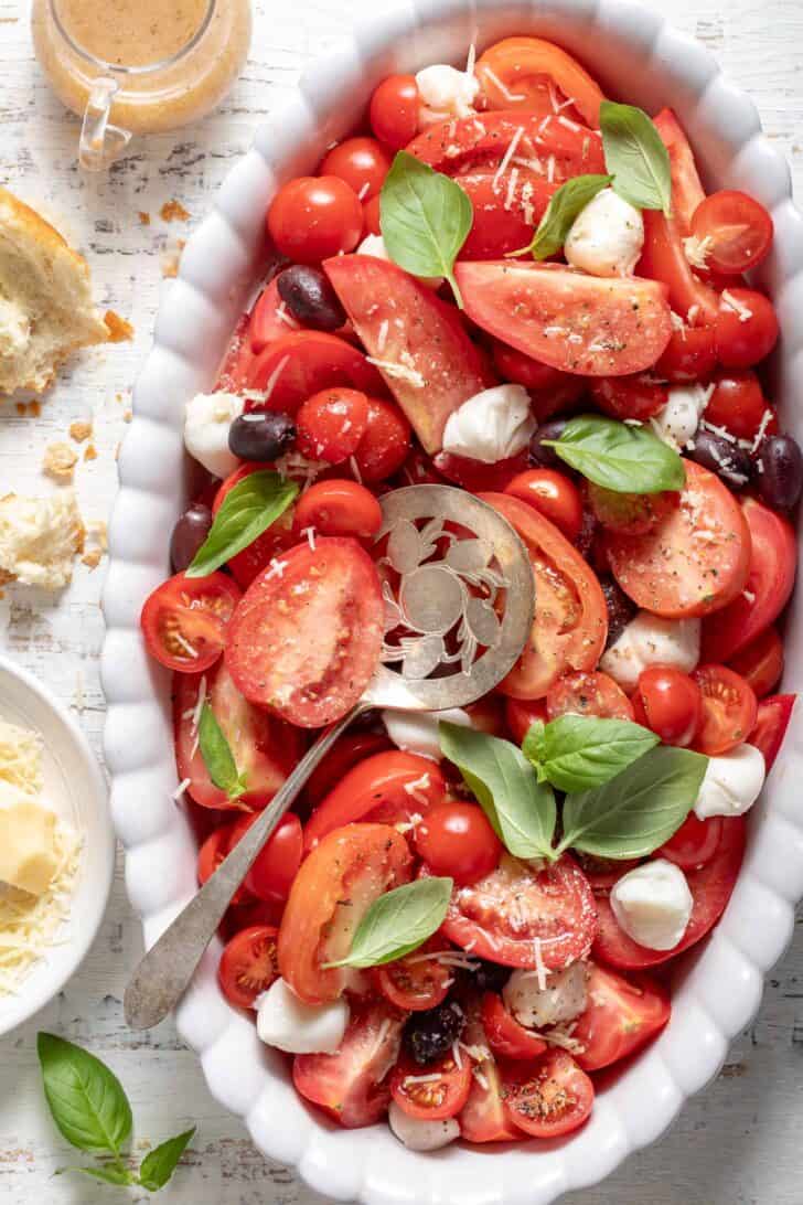 A white oval platter filled with Caprese Salad (sliced tomatoes with cheese balls, olives and fresh basil leaves).