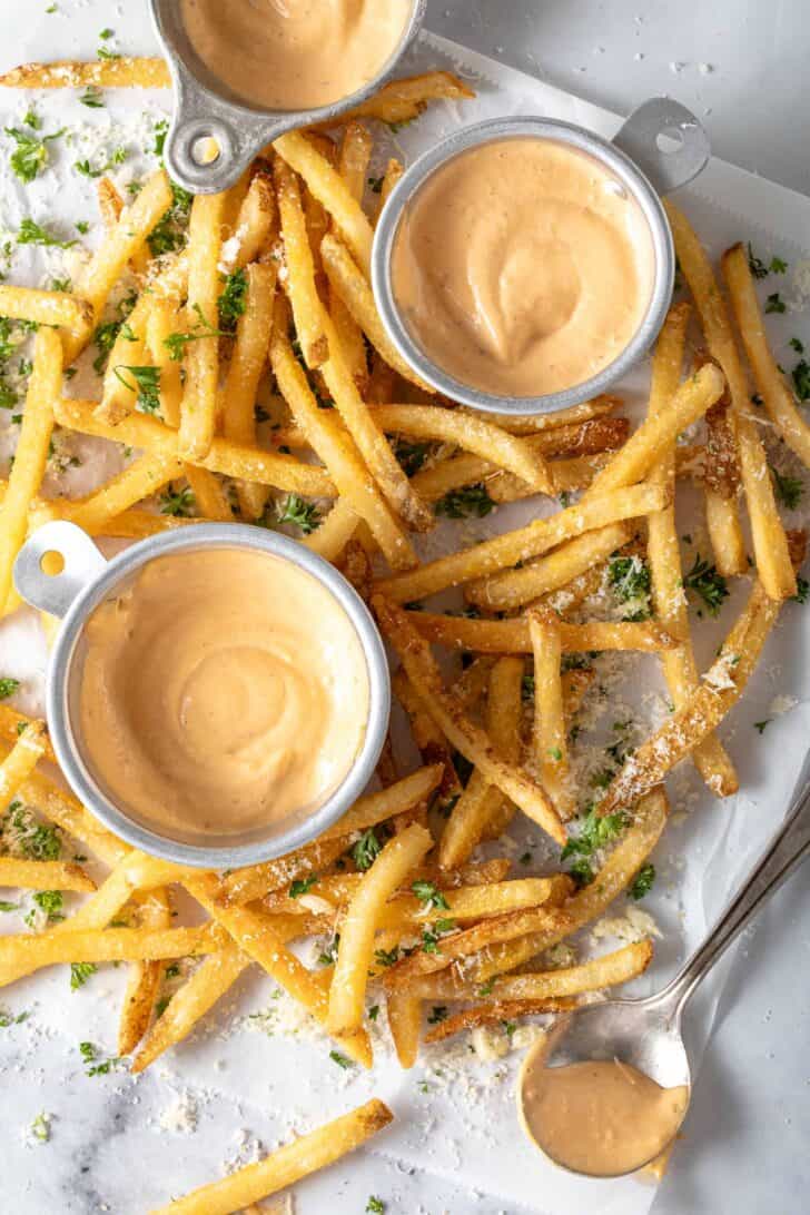 French fries are scattered on a white paper. Three cups filled with a spicy sauce sits around the French fries.