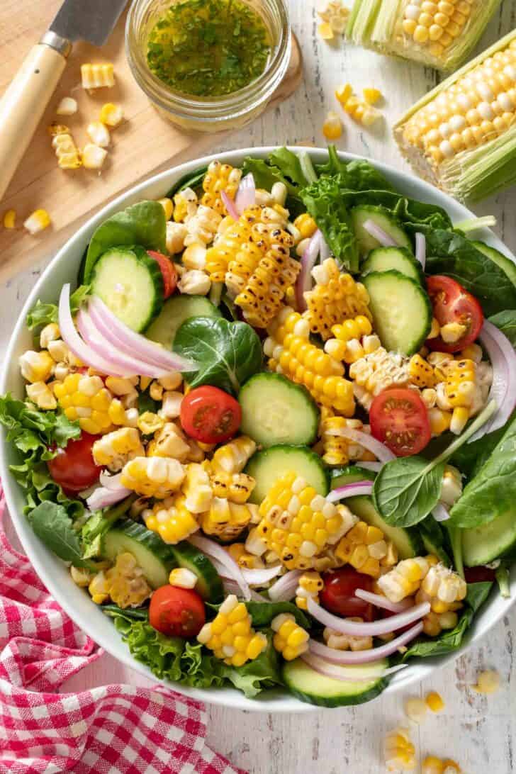 A white bowl filled with fresh Summer Corn Salad filled with corn, cucumber slices, spinach leaves, red onion and cherry tomatoes. A red checkered napkin sits next to the bowl.