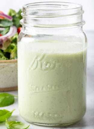 A clear glass mason jar filled with Creamy Basil Salad Dressing. A tan colored bowl filled with salad sits next to the jar.