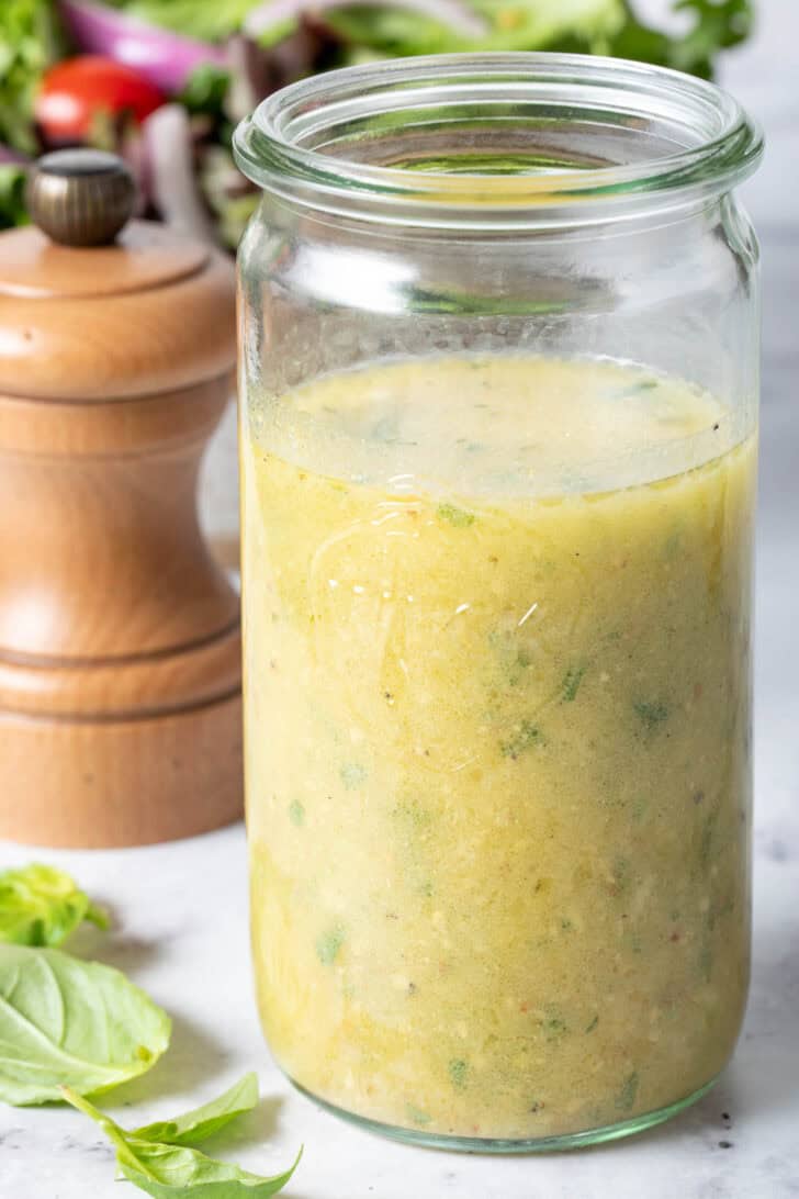 A clear glass mason jar filled with Fresh Basil Vinaigrette sits on a marble countertop. A wooden pepper mill sits next to the jar.