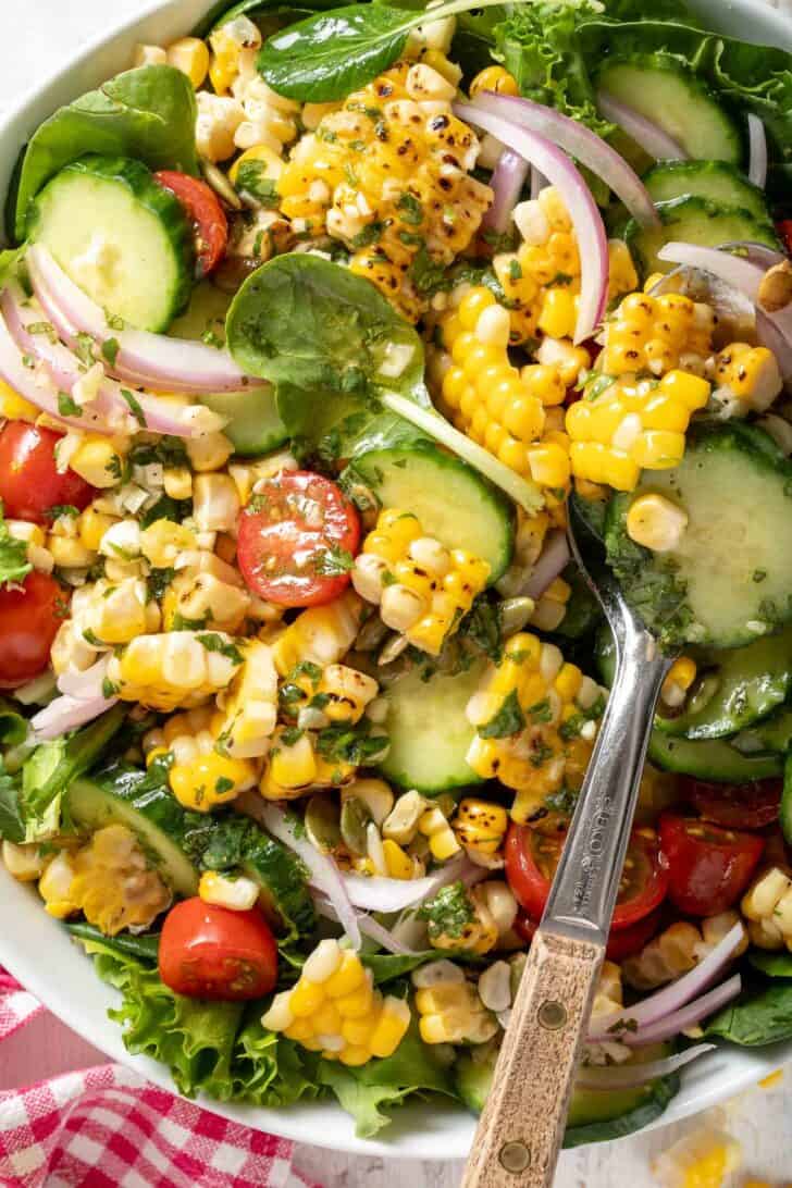 A white bowl filled with Summer Corn Salad made with grilled corn, spring mix, tomatoes, onion, cucumber and pepitas. A spoon with a wooden handle rests in the bowl.