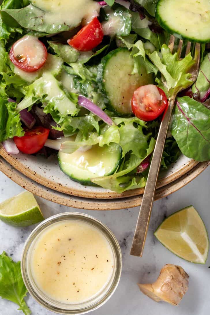 A tan colored bowl filled with mixed greens, cherry tomatoes and cucumber slices. A small glass bowl filled with dressing sits next to the bowl.