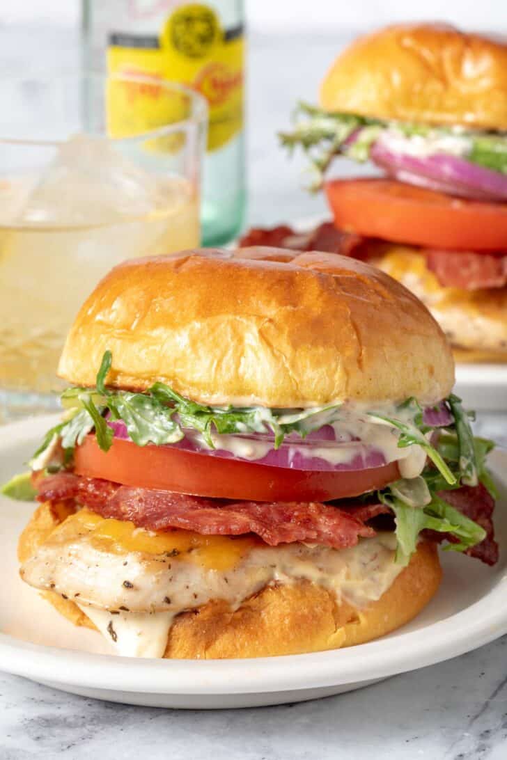Two grilled chicken sandwiches on white plates. A glass filled with a beverage sits next to the sandwiches.