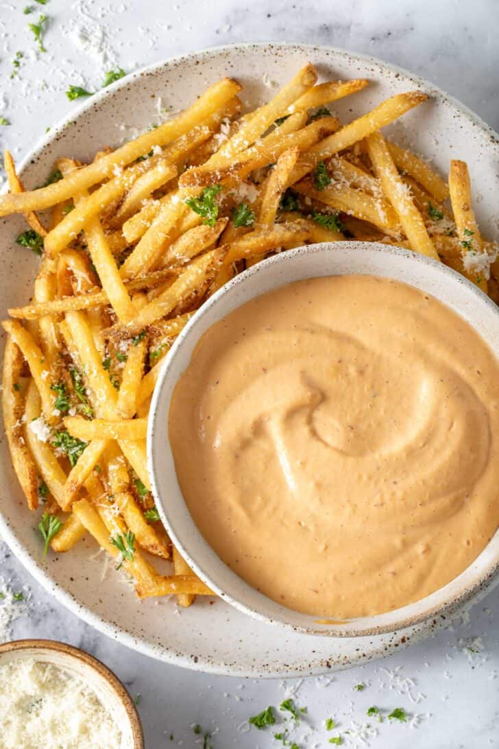 A bowl filled with a spicy sauce sits on a plate filled with French fries.