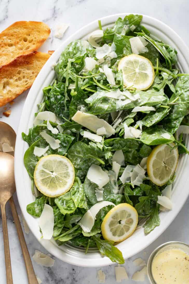 A white oval platter filled with Parmesan Salad. Golden salad serves sit next to the platter - for a simple arugula salad recipe.