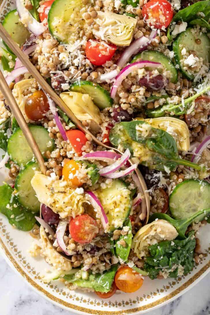 A white platter filled with mixed greens, cherry tomatoes, red onion, cucumbers, artichoke hearts and kalamata olives. Gold colored salad servers rest on the platter.