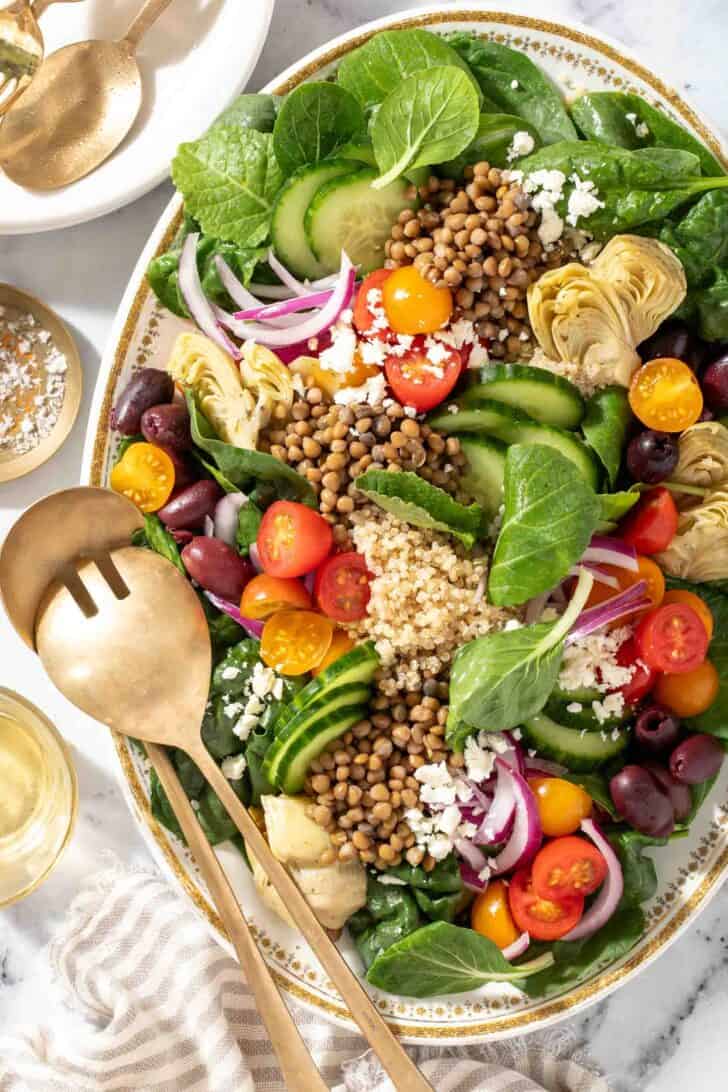 A white platter filled with Lentil Quinoa salad made with lentils, quinoa, spinach, cherry tomatoes, red onion, cucumbers, artichoke hearts and kalamata olives. Gold colored salad servers rest on the platter.