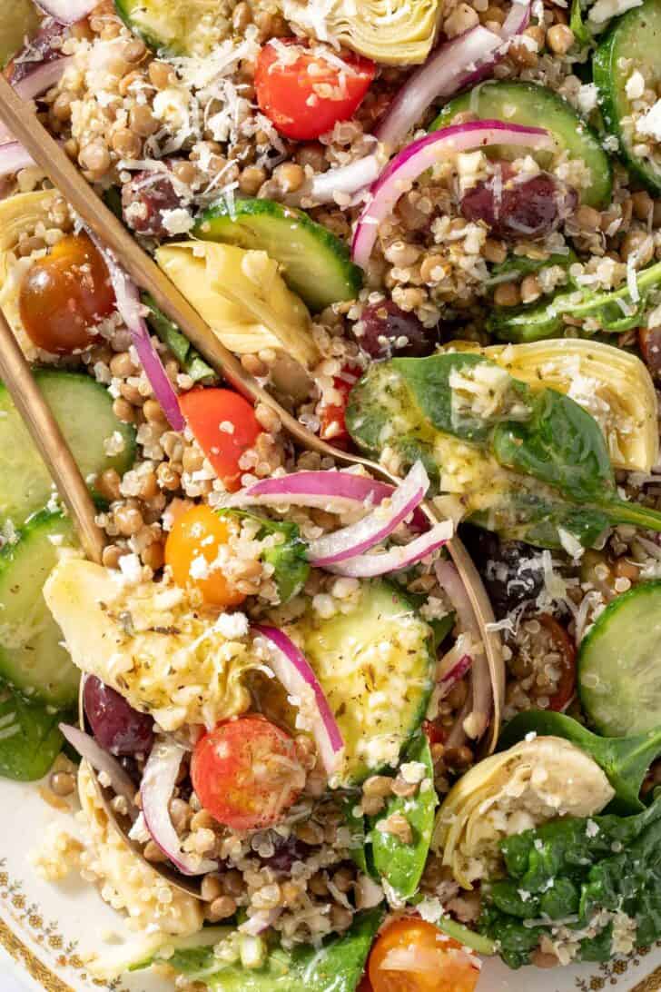 A white platter filled with mixed greens, cherry tomatoes, red onion, cucumbers, artichoke hearts and kalamata olives. Gold colored salad servers rest on the platter.