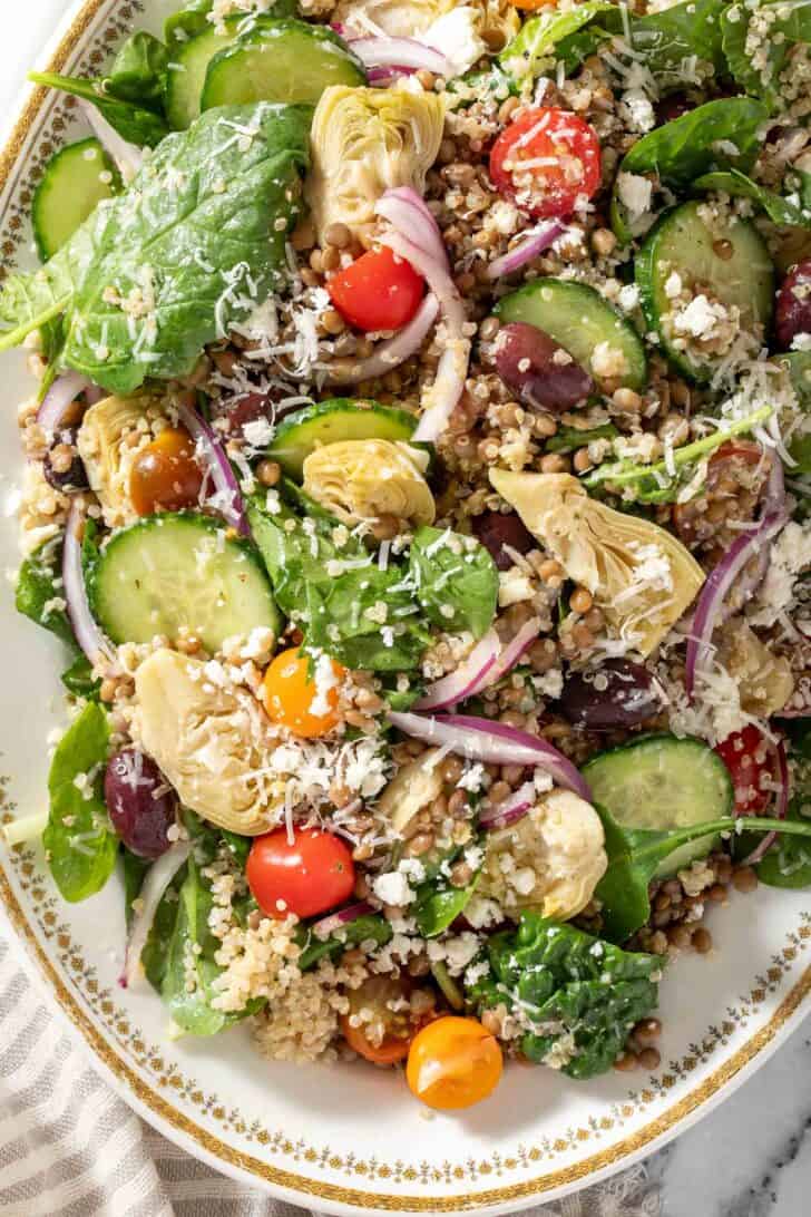 A white platter filled with spinach, cherry tomatoes, red onion, cucumbers, artichoke hearts and kalamata olives. Gold colored salad servers rest on the platter.