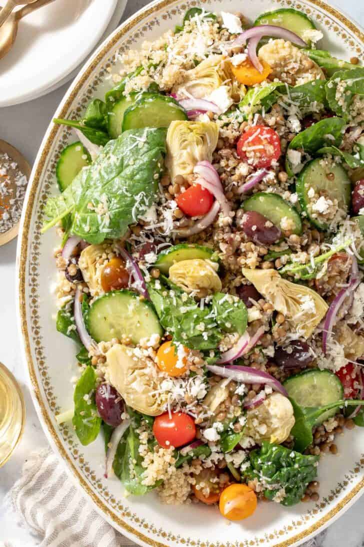 A white platter filled with Lentil Quinoa salad made with lentils, quinoa, spinach, cherry tomatoes, red onion, cucumbers, artichoke hearts and kalamata olives. Gold colored salad servers rest on the platter.